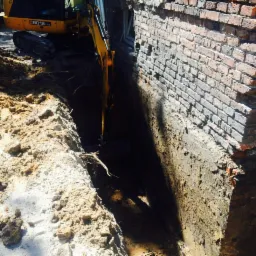 Żółta mini koparka w trakcie wykonywania wąskiego wykopu wzdłuż ceglanego fundamentu budynku, widoczne nierówne ściany wykopu i sterta ziemi obok.