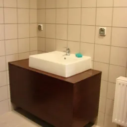 Biała, prostokątna umywalka nablatowa na ciemnobrązowej szafce w łazience z jasnymi, kwadratowymi płytkami i grzejnikiem żeberkowym z boku.