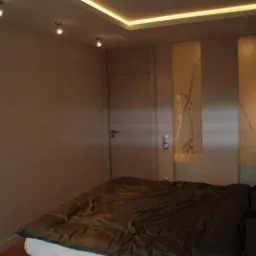 Wykończona sypialnia z podświetlanym sufitem, beżowymi ścianami i dekoracyjnymi panelami na ścianie za łóżkiem. Widoczne oświetlenie punktowe i minimalistyczny design.