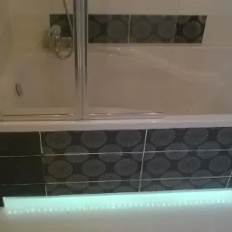 Biała wanna z parawanem nawannowym, obudowana czarnymi płytkami z szarym wzorem w koła, podświetlona listwą LED.