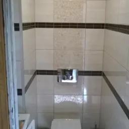 Wnętrze wykończonej łazienki z jasnymi płytkami z dekoracyjnym wzorem, ciemnym pasem na ścianach i czarną podłogą, widoczna toaleta, umywalka oraz akcesoria łazienkowe.