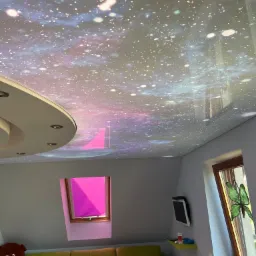 Sufit napinany z motywem kosmosu w pokoju dziecięcym, widoczne zabawki i okno dachowe z różową roletą.