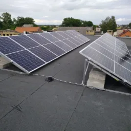 Dwa rzędy paneli fotowoltaicznych zamontowane na płaskim dachu pokrytym papą, widok z góry, w tle zabudowa jednorodzinna i pochmurne niebo.