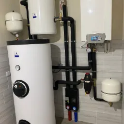 Biały, pionowy zasobnik ciepłej wody użytkowej De Dietrich z czarnym panelem sterowania, połączony rurami z wiszącym kotłem gazowym tej samej marki, instalacja w pomieszczeniu wyłożonym płytkami...