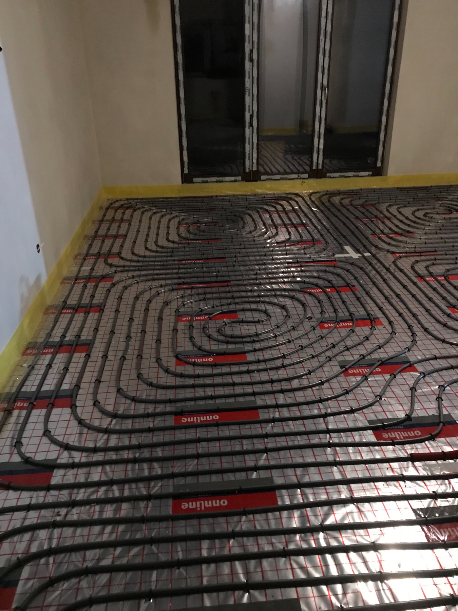 Rozłożone rury ogrzewania podłogowego na folii izolacyjnej z widocznymi oknami w tle.