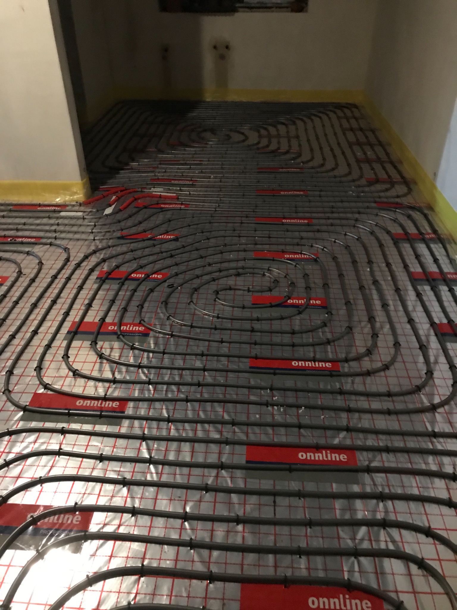 Rozłożone rury ogrzewania podłogowego na folii odblaskowej z siatką, widoczne czerwone elementy mocujące rury, żółta taśma na ścianach.