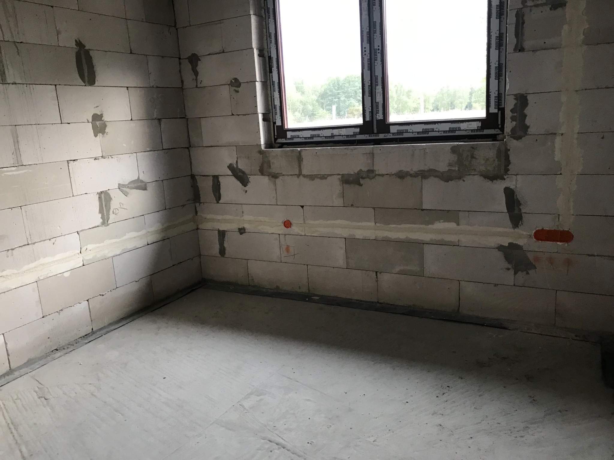 Surowe wnętrze pomieszczenia w trakcie prac wykończeniowych: ściany z betonu komórkowego z widocznymi zaprawami, wstawione okno z profili Rehau, w ścianie widoczne otwory pod instalacje elektryczne.