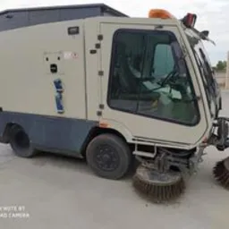Beżowa zamiatarka przemysłowa z kabiną, widoczna z boku na betonowym placu, z obrotowymi szczotkami z przodu. Pomarańczowe światło ostrzegawcze na dachu. W tle budynek gospodarczy.