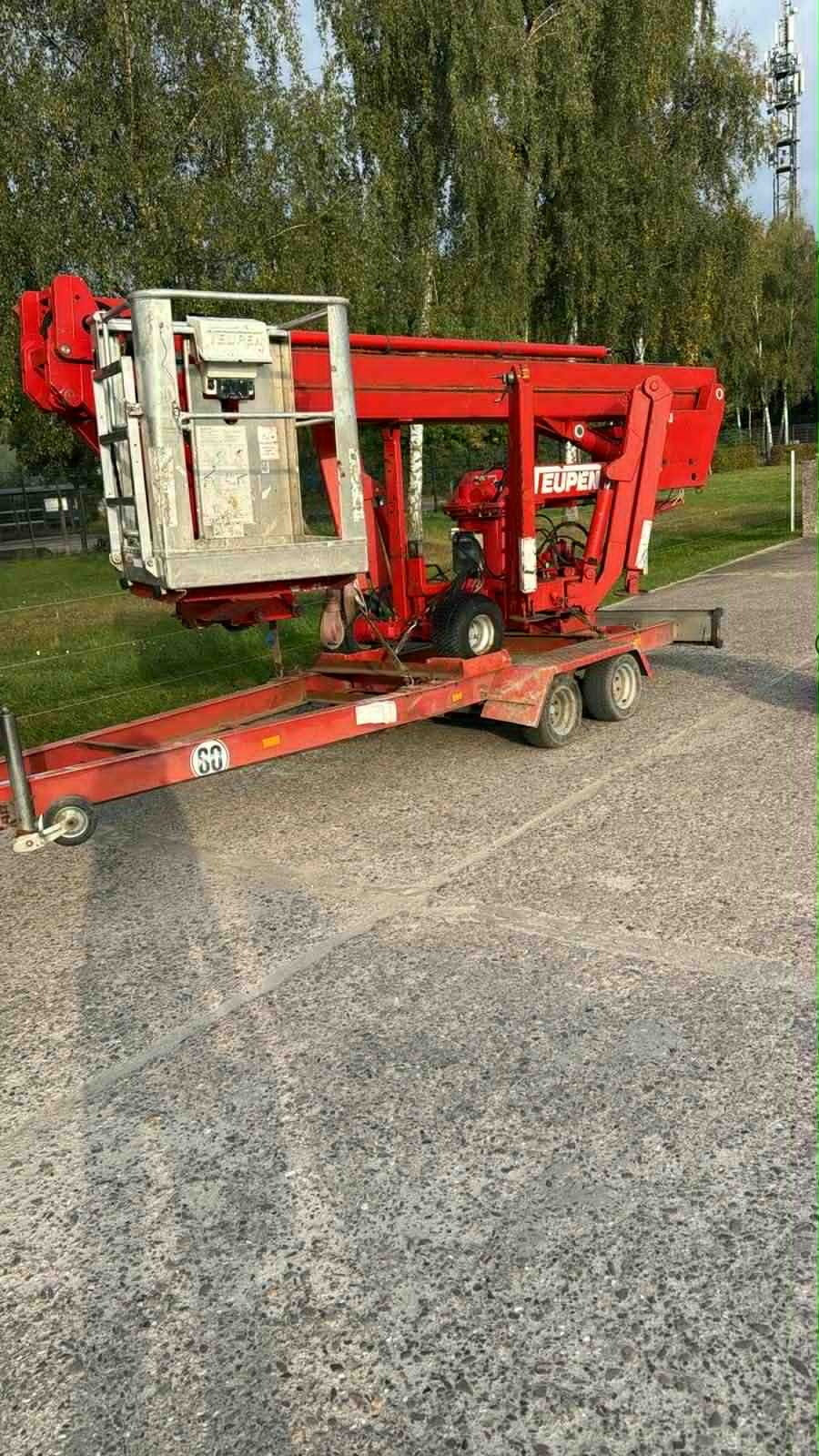 Czerwony podnośnik koszowy na przyczepie, z widocznym koszem i ramieniem, zaparkowany na utwardzonej powierzchni. W tle drzewa i słup telekomunikacyjny.