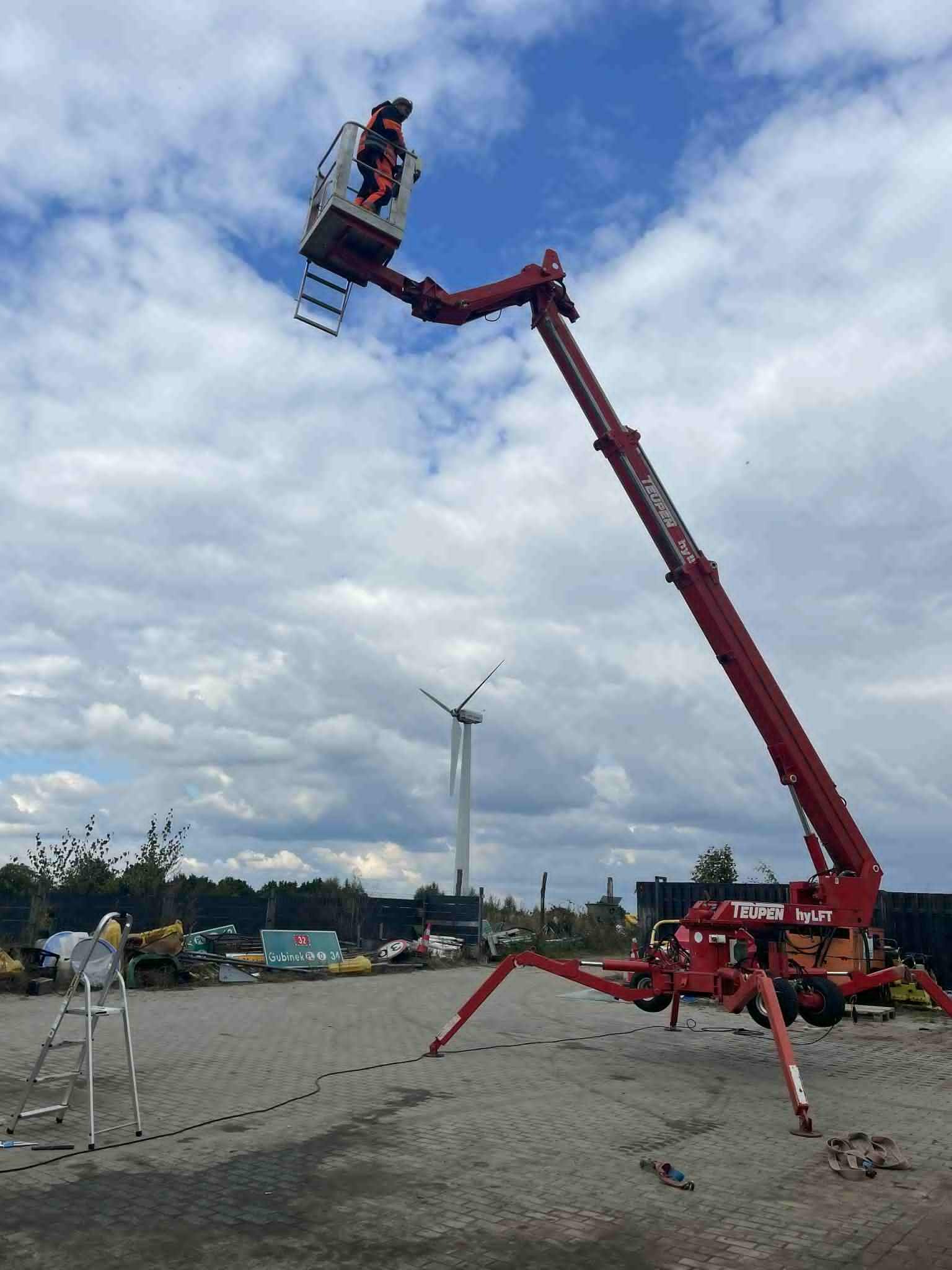 Czerwony podnośnik koszowy Teupen hyLIFT z operatorem na wysokości, w tle wiatrak i pochmurne niebo. Na ziemi drabina i narzędzia.
