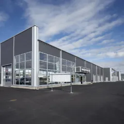 Nowoczesna hala stalowa z szarymi panelami i dużymi oknami, widok z zewnątrz na tle błękitnego nieba z chmurami. Widoczny napis firmowy nad wejściem.