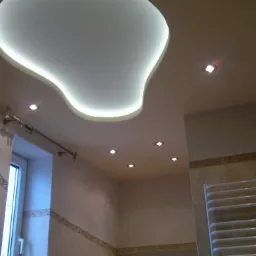 Wykończona łazienka w beżowych odcieniach z podwieszanym sufitem o nieregularnym kształcie, podświetlonym ledami. Na ścianach płytki z delikatnym wzorem, grzejnik drabinkowy.