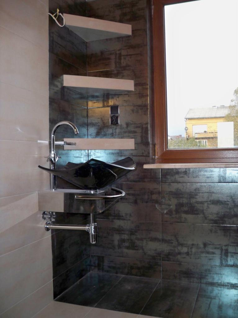 Nowoczesna łazienka z czarną, szklaną umywalką na tle ściany z płytek imitujących metal. Widoczne drewniane półki i okno z widokiem na budynek.