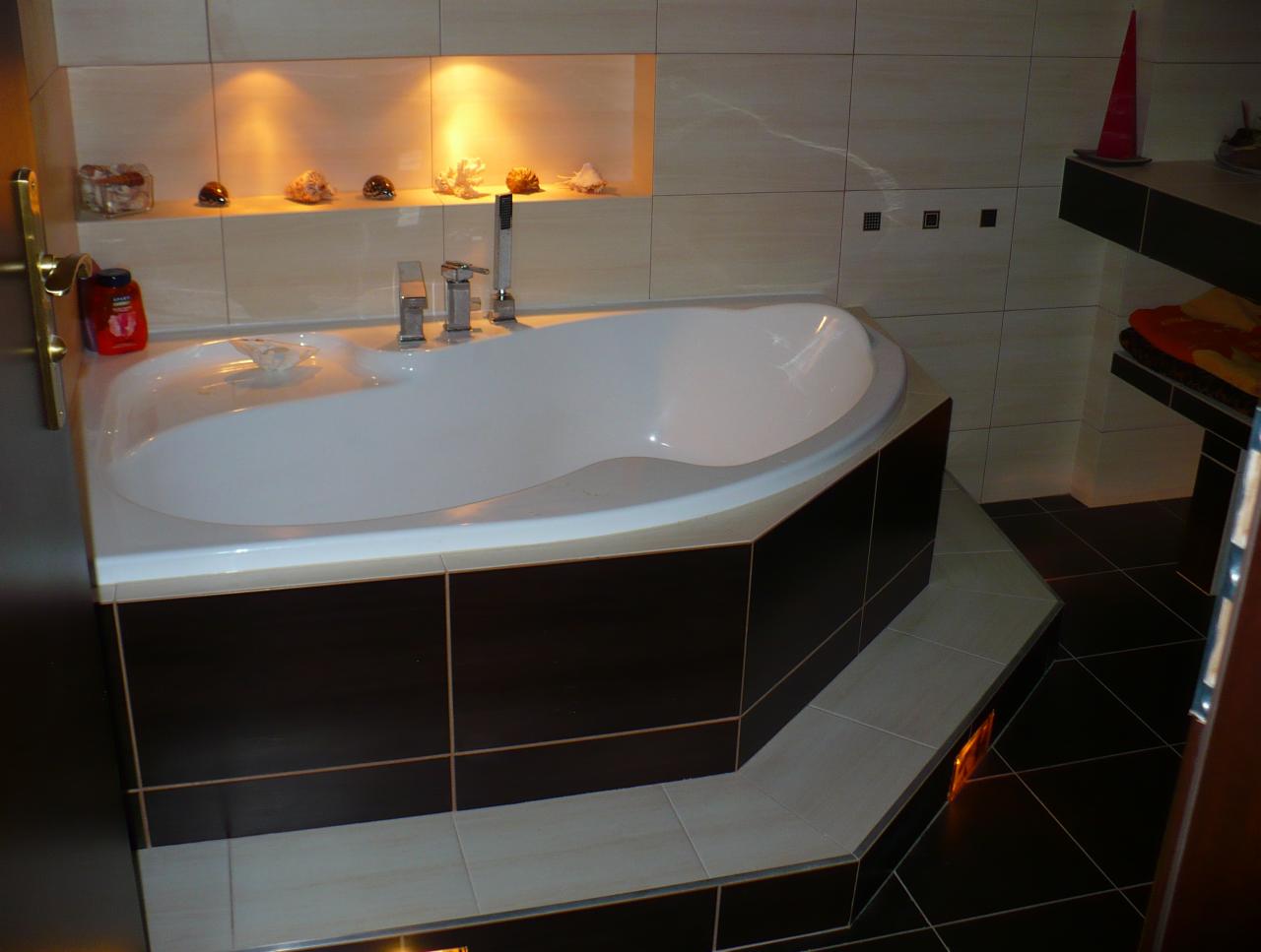 Nowoczesna łazienka z asymetryczną wanną obudowaną ciemnymi płytkami i podświetleniem. Na półce dekoracje z muszelek, a na ścianach beżowe płytki.