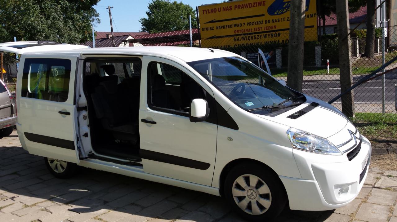 Biały Citroen SpaceTourer z otwartymi drzwiami suwanymi, zaparkowany na chodniku, z widocznym fragmentem reklamy w tle. Dostępny do wynajmu.