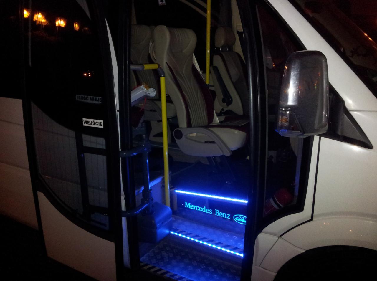 Wnętrze białego busa Mercedes-Benz z oświetleniem LED na schodkach wejściowych. Widoczne fotele pasażerskie i elementy wyposażenia. Nocne oświetlenie zewnętrzne.