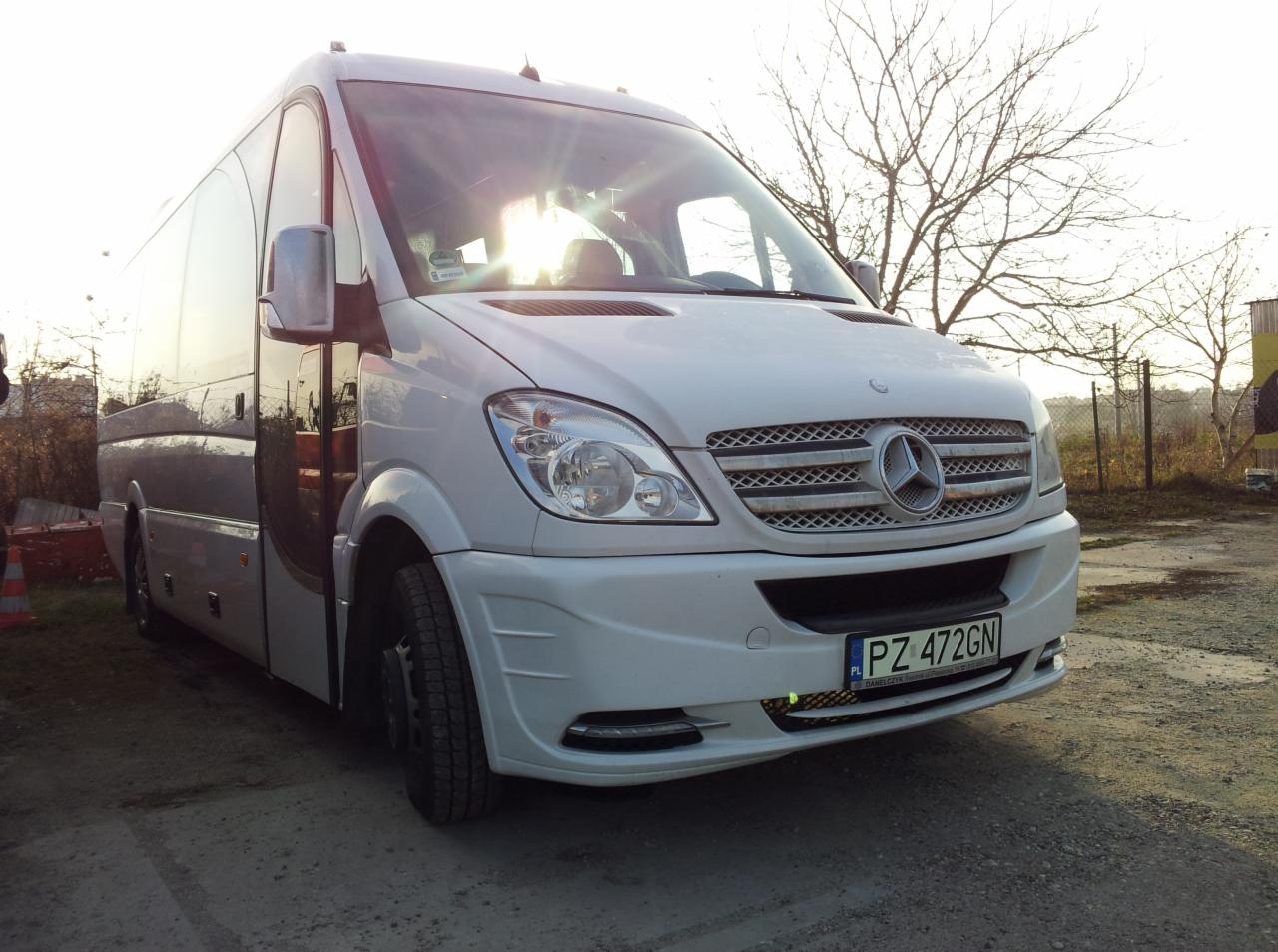 Biały Mercedes Sprinter z widoczną tablicą rejestracyjną PZ 472GN, zaparkowany na żwirowym podłożu w słoneczny dzień. Widoczne drzewa w tle.