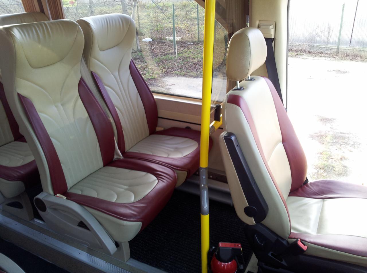 Wnętrze autobusu z beżowo-bordowymi skórzanymi fotelami, żółtą poręczą i gaśnicą. Widok na okno i krajobraz zewnętrzny. Komfortowe siedzenia w pojeździe.