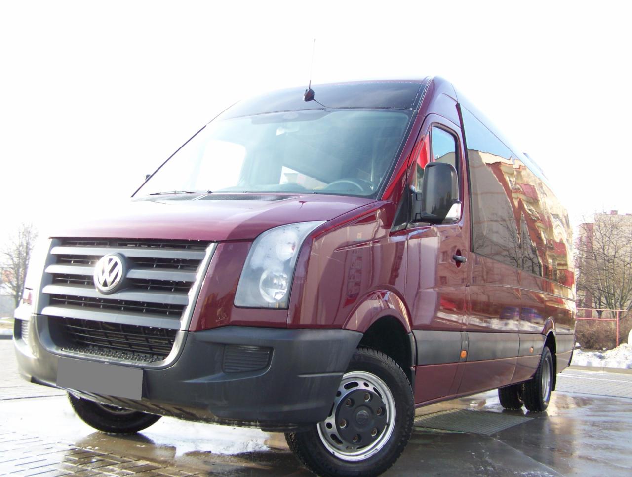 Czerwony bus Volkswagen Crafter, widok z przodu, zaparkowany na mokrej powierzchni. W tle budynek odbijający się w szybach.