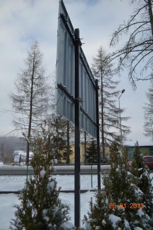 Tył metalowej konstrukcji tablicy reklamowej na zewnątrz w zimowy dzień, widoczne drzewa bez liści i iglaki pokryte śniegiem, data 26.01.2013 w prawym dolnym rogu.