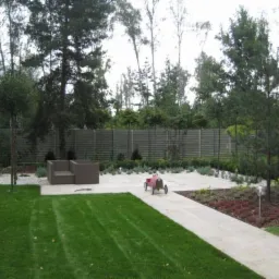 Zadbany ogr&oacute;d z r&oacute;wno przystrzyżonym trawnikiem, nowoczesnym meblem ogrodowym i dekoracyjnym żwirkiem. Na ścieżce stoi czerwony w&oacute;zek.