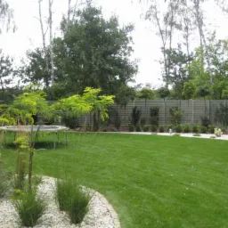 Zadbany trawnik z trampoliną i ozdobnymi roślinami na tle drewnianego ogrodzenia. Widok na zielony ogród z elementami dekoracyjnymi.