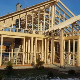 Surowa konstrukcja drewniana domu jednorodzinnego w trakcie budowy, widoczne elementy szkieletu i więźby dachowej, drabina oparta o ścianę, w tle inne budynki i drzewa.