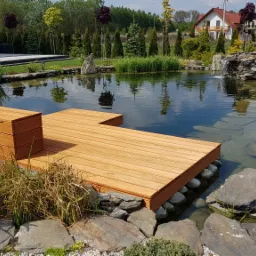 Drewniany pomost z siedziskiem nad stawem w ogrodzie, otoczony kamieniami i roślinnością, z widokiem na mały wodospad i domy w tle.