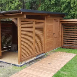 Drewniana altana z lamelowymi ścianami i ścieżką z desek na trawniku, widoczny fragment dachu i rynny, w tle drzewa.