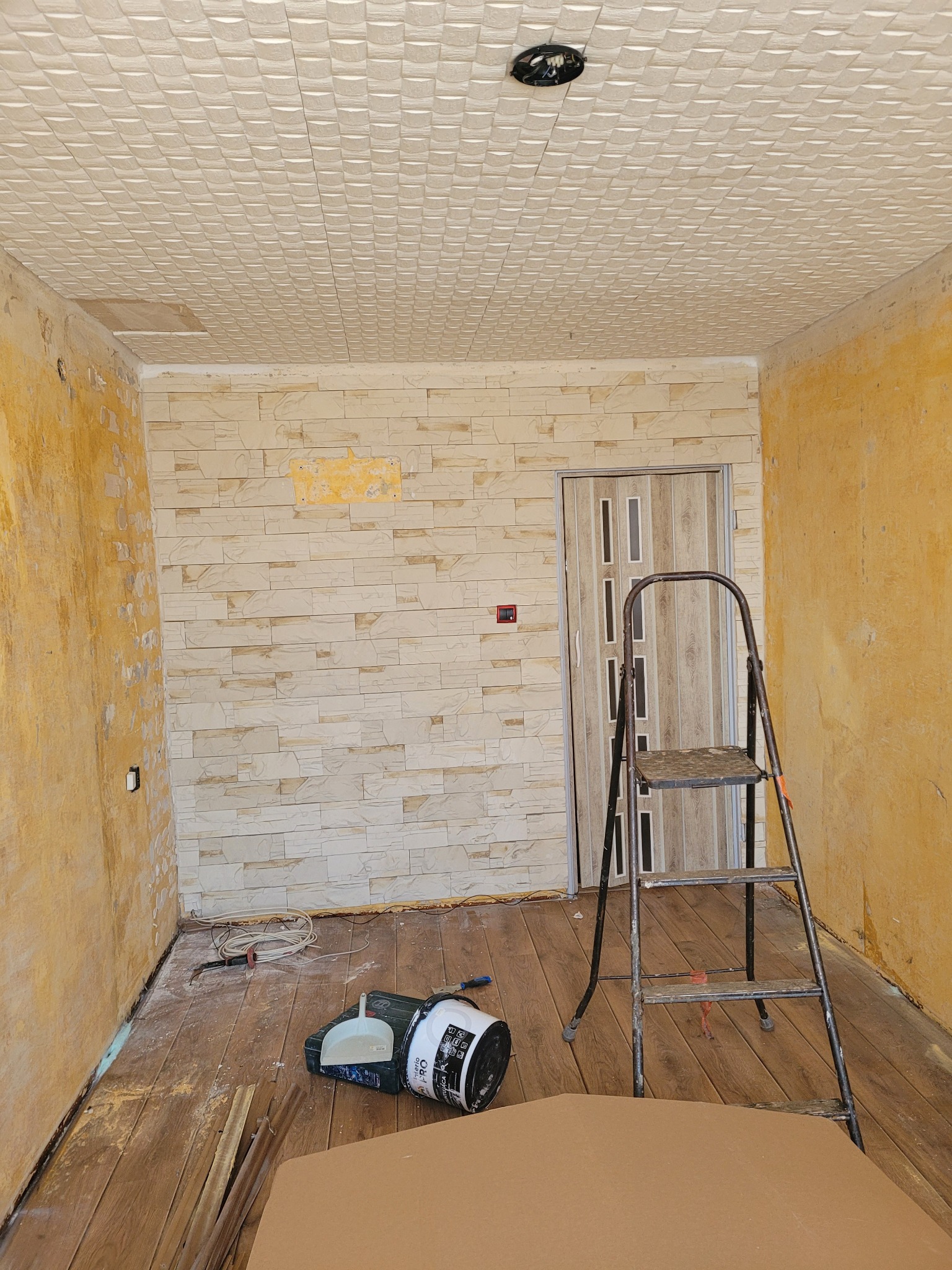 Wnętrze w trakcie remontu: ściana wyłożona dekoracyjną cegłą, sufit z kasetonami, drabina, wiadro z farbą i narzędzia na podłodze. Żółte ściany w tle.