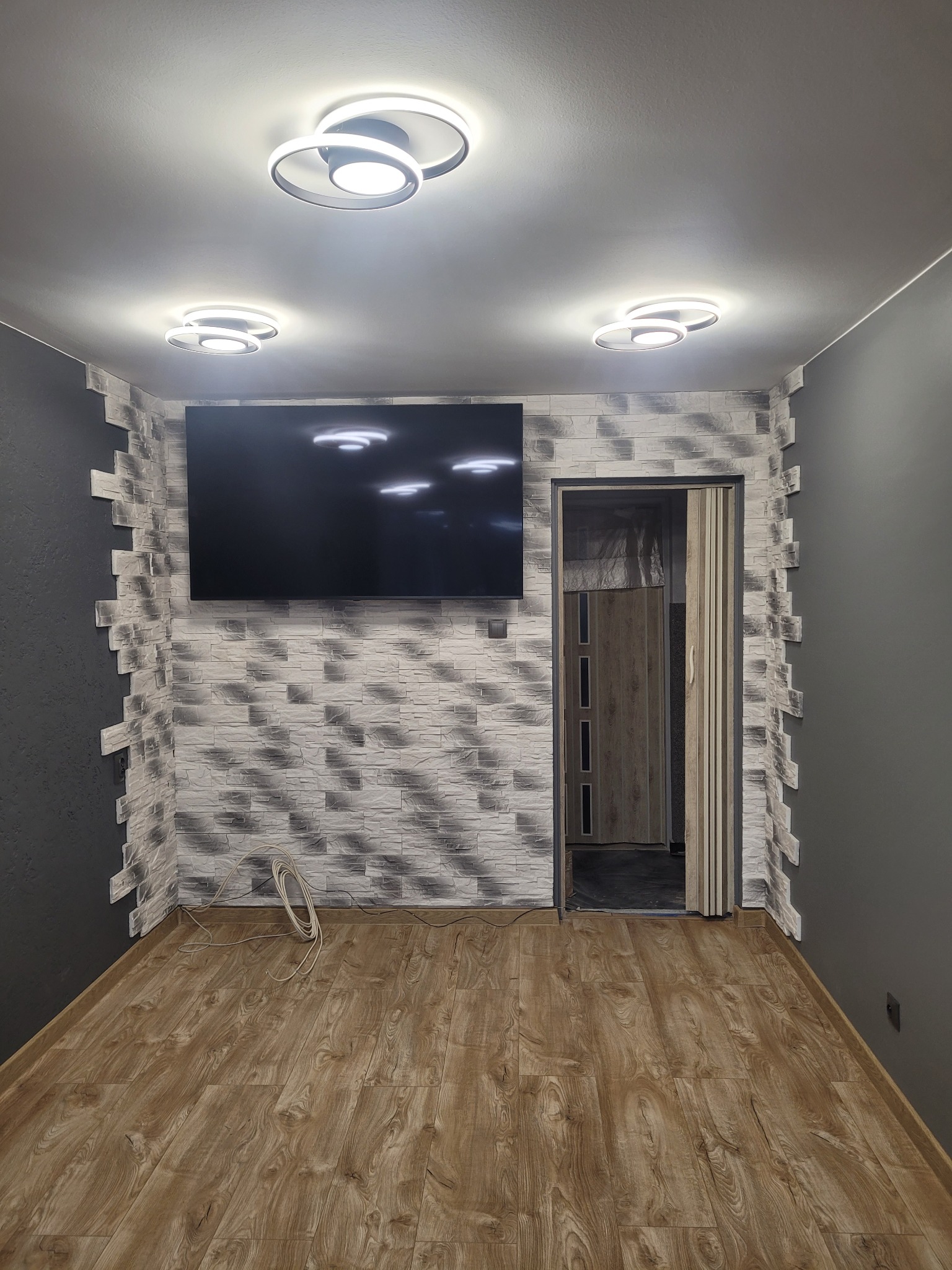 Pokój po remoncie: panele podłogowe, ściany w szarościach z dekoracyjną cegłą, nowoczesne oświetlenie sufitowe, wiszący telewizor i otwarte drzwi.