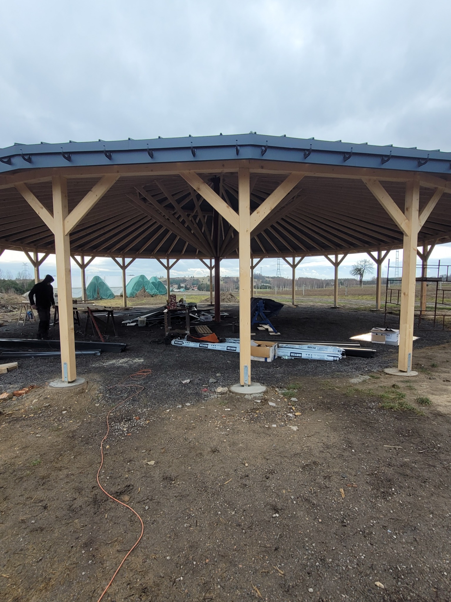 Budowa altany: drewniana konstrukcja z niebieskim dachem na żwirowym podłożu. Widoczne materiały budowlane i osoba pracująca w tle. Pochmurne niebo.