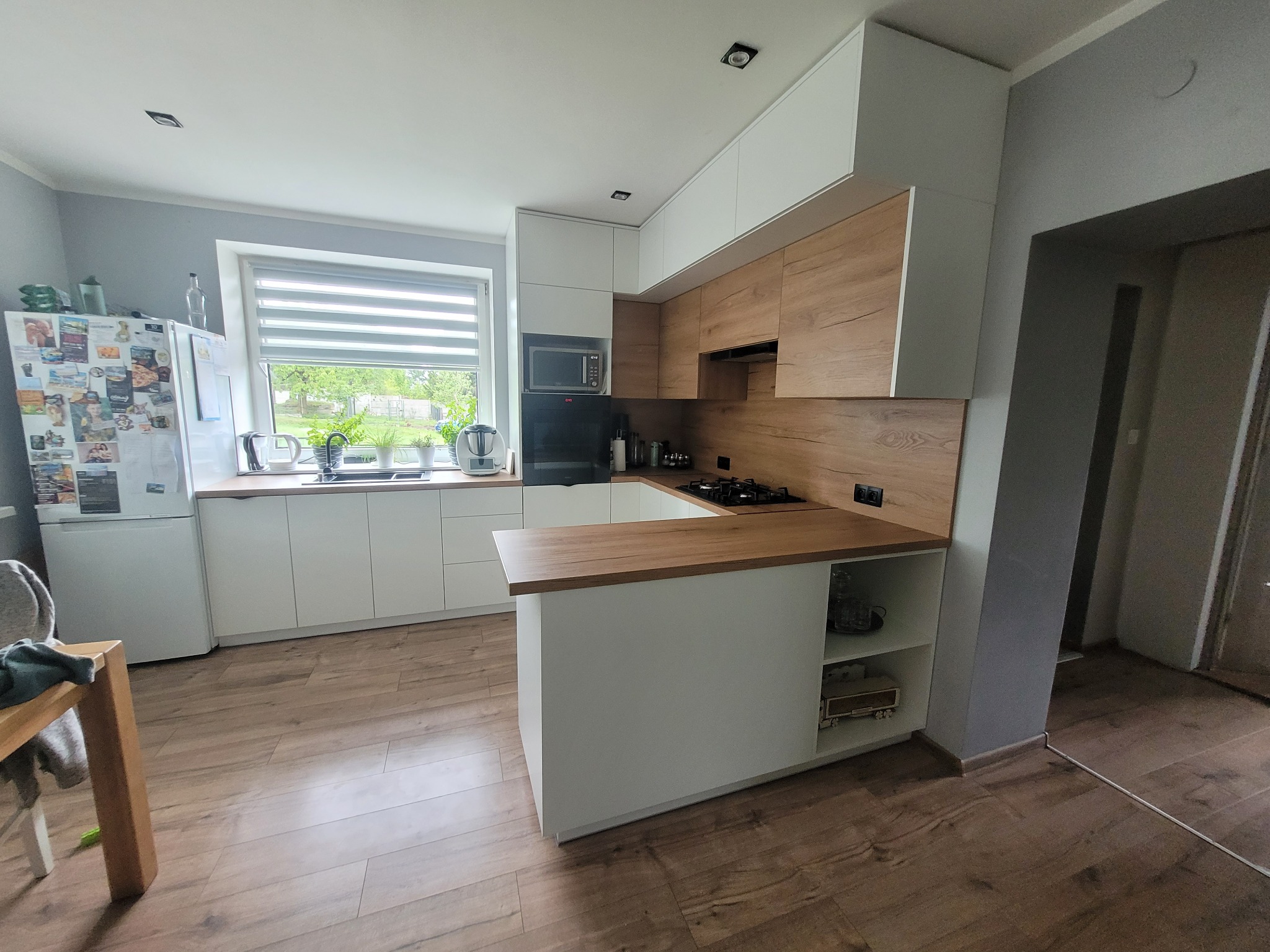 Nowoczesna kuchnia z białymi szafkami i drewnianymi akcentami, widok na okno z roletami. Jasne wnętrze z drewnianą podłogą i wyspą kuchenną.