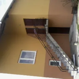 Dwukolorowa elewacja budynku z zewnętrznymi schodami z ozdobną, metalową balustradą prowadzącymi do drzwi na piętrze. Widoczny fragment ogrodu i nieba.