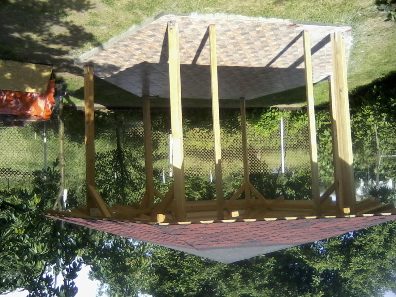 Konstrukcja drewnianej altany ogrodowej z czerwonym dachem i podłogą z płytek, w trakcie budowy we Włocławku, otoczona zielenią.