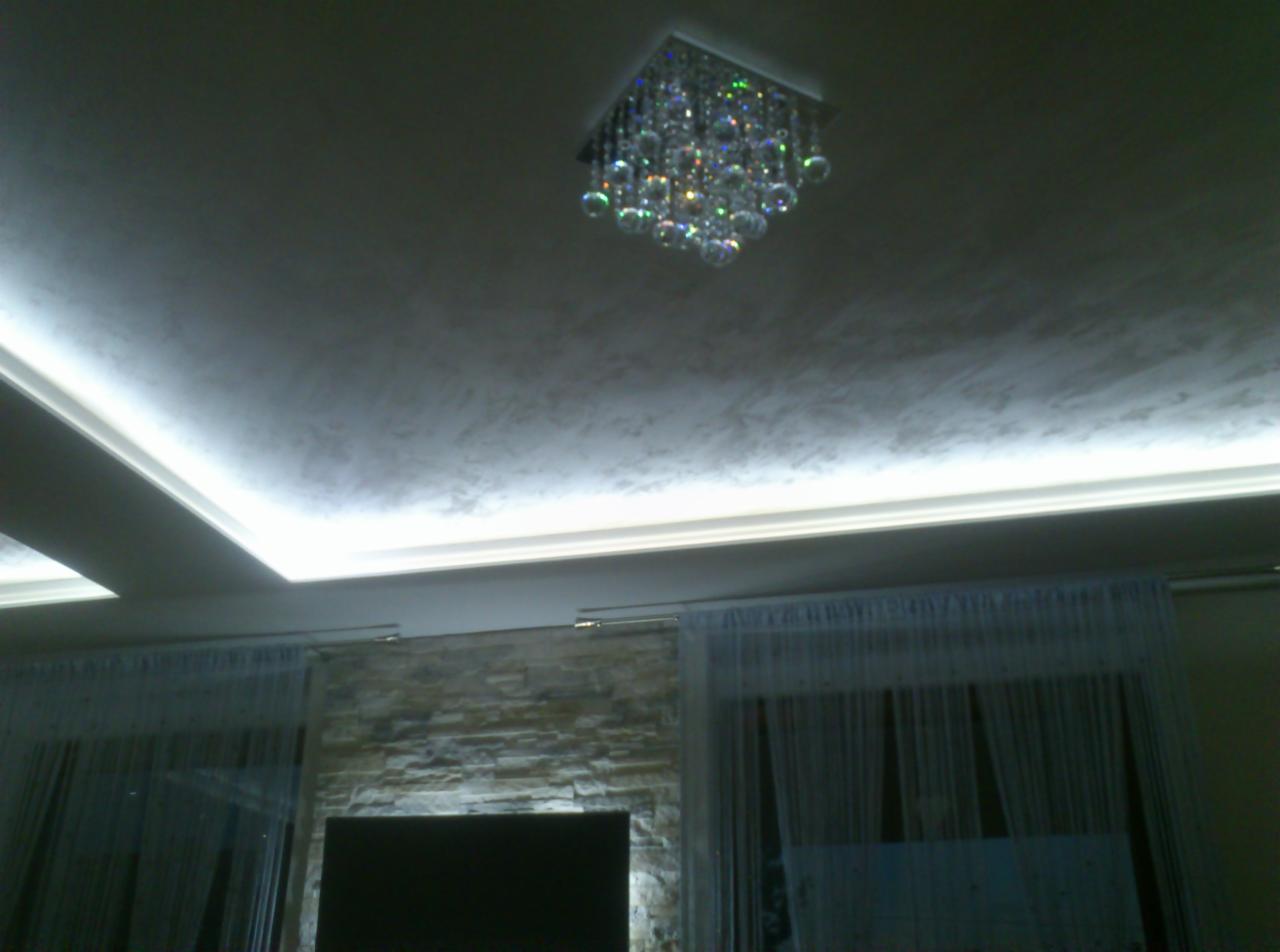 Wykończony sufit z dekoracyjną strukturą i oświetleniem LED wzdłuż krawędzi, kryształowy żyrandol w centrum, fragment ściany z kamienia dekoracyjnego i firany w tle.