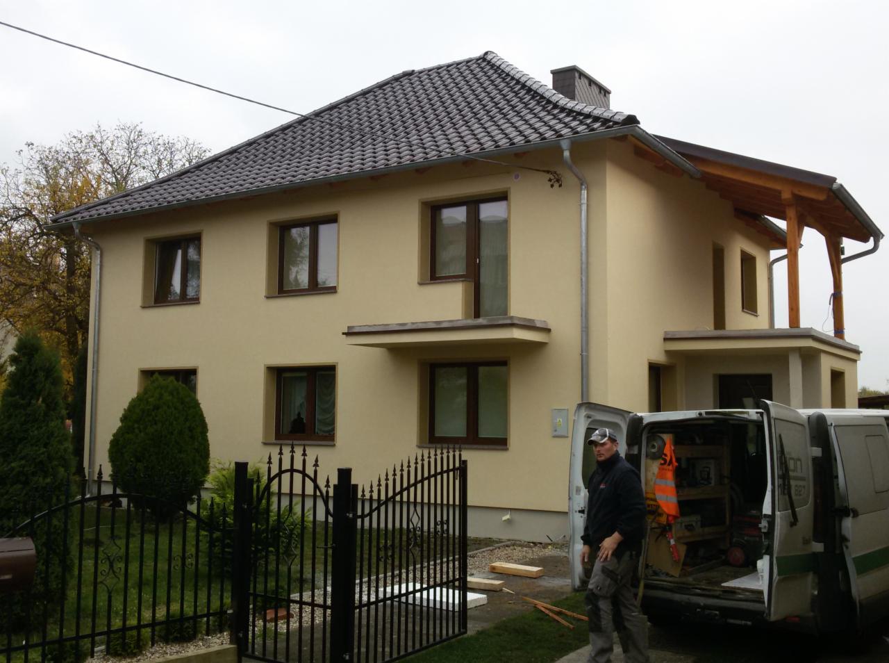 Dwukondygnacyjny dom z beżową elewacją, ciemną dachówką i drewnianą konstrukcją balkonu, widoczny przez czarne, metalowe ogrodzenie, z pracownikiem budowlanym i otwartym busem pełnym narzędzi.