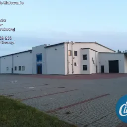 Biały budynek magazynowy lub hali produkcyjnej po czyszczeniu elewacji, widoczny duży parking z kostki brukowej i logo firmy Tom Clean w rogu