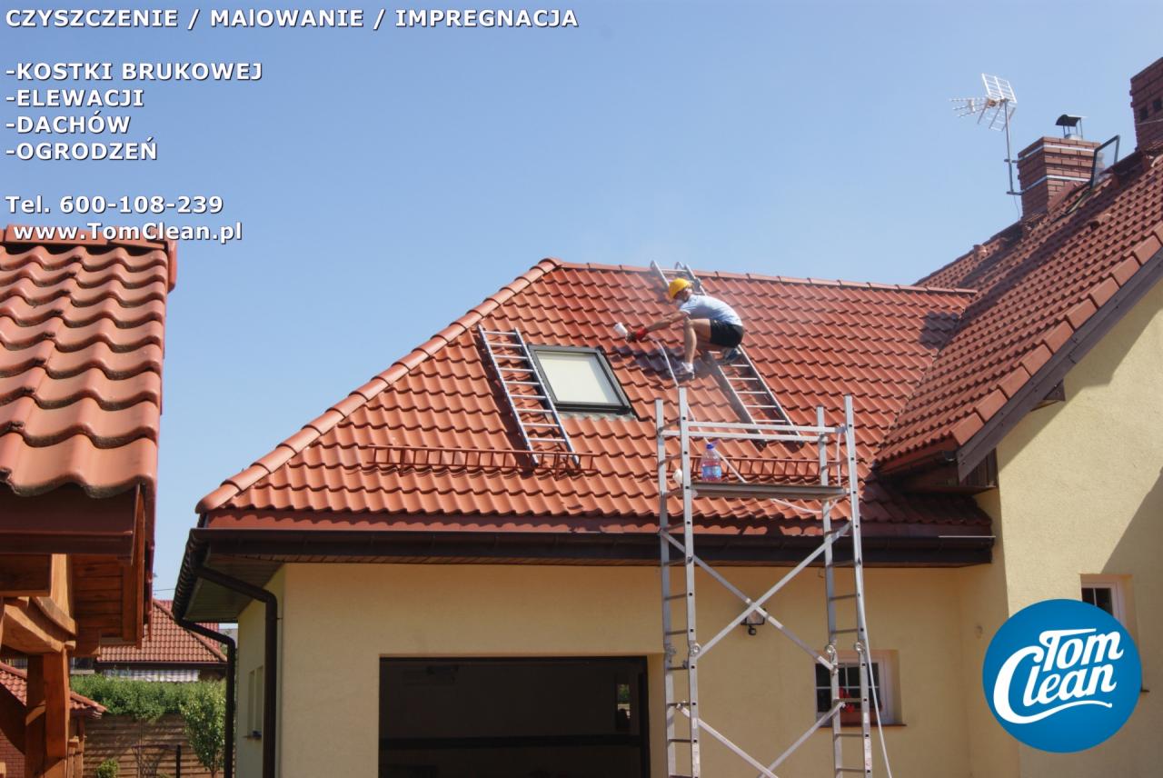 Pracownik w kasku, na drabinie, czyści dach z czerwonej dachówki w Białymstoku, używając myjki ciśnieniowej, obok widoczny fragment elewacji domu.