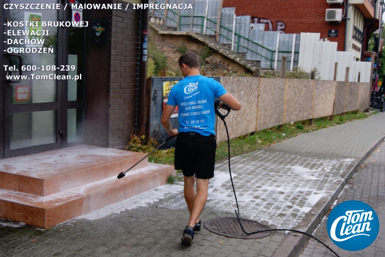Mężczyzna w niebieskiej koszulce z logo Tom Clean czyści schody wejściowe do budynku za pomocą myjki ciśnieniowej, w tle widoczna ulica i budynek z cegły.