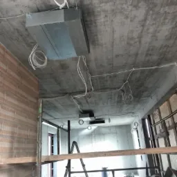 Montaż centrali wentylacyjnej pod betonowym sufitem w surowym stanie, widoczne przewody i elementy instalacji, rusztowanie w tle.