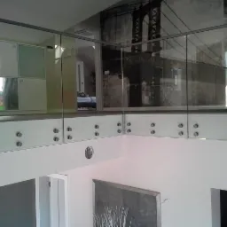 Szklana balustrada z metalowymi elementami mocującymi w nowoczesnym wnętrzu. W tle widoczna fototapeta z motywem mostu.