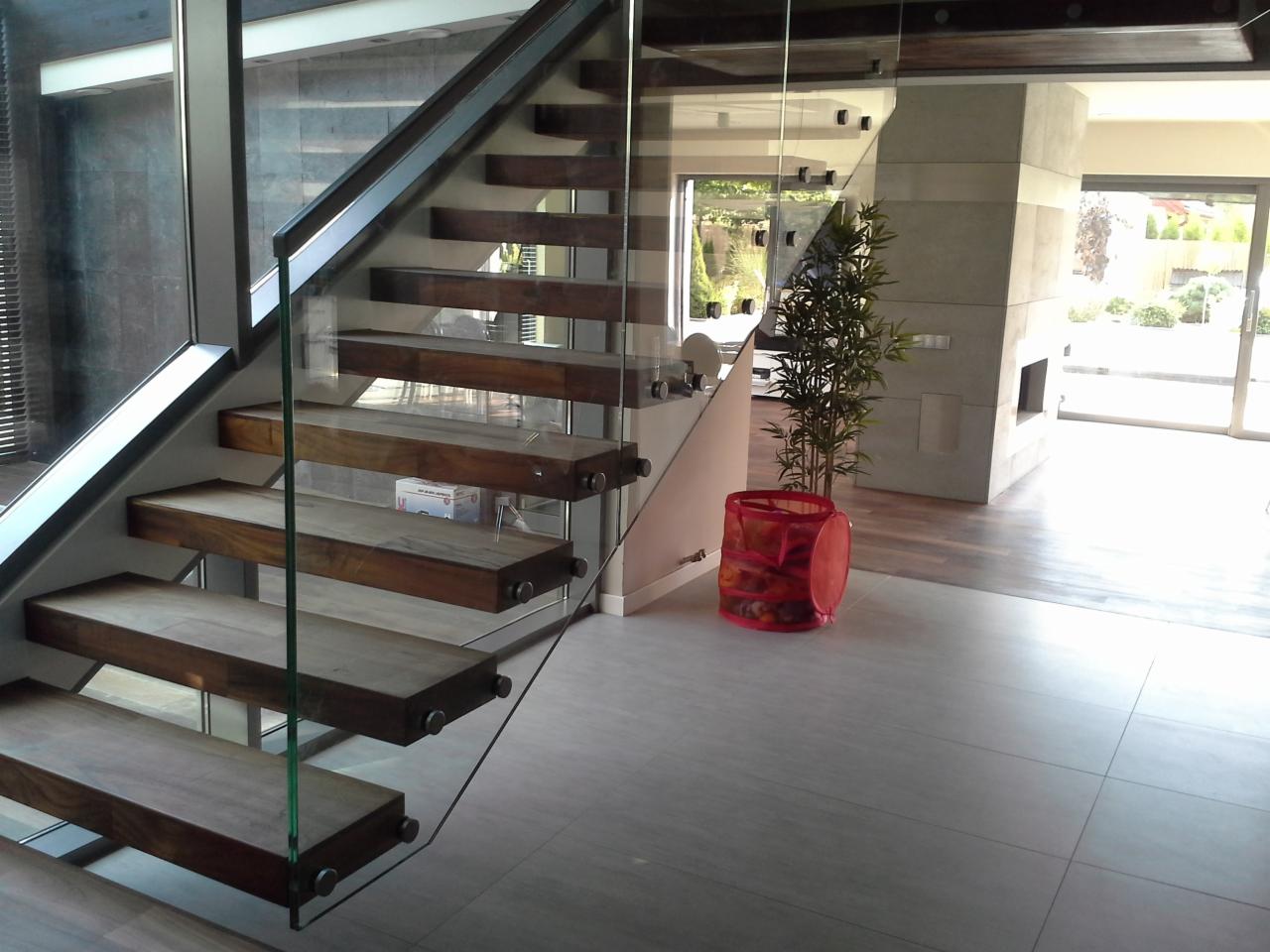 Nowoczesne schody z drewnianymi stopniami i szklaną balustradą w jasnym wnętrzu. Widoczne detale mocowania szkła i minimalistyczny design.