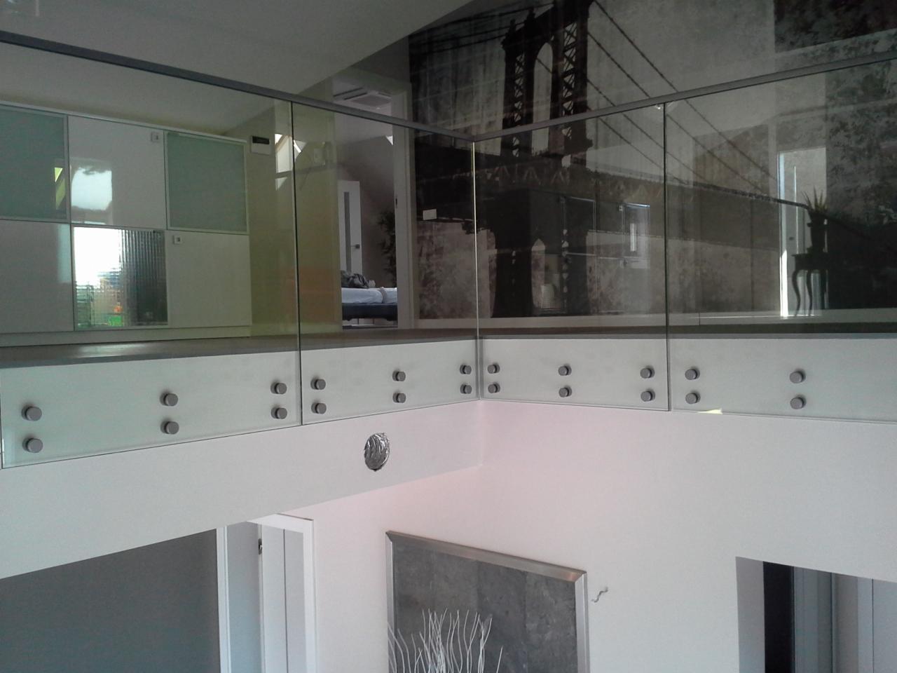 Szklana balustrada z metalowymi elementami mocującymi w nowoczesnym wnętrzu. W tle widoczna fototapeta z motywem mostu.