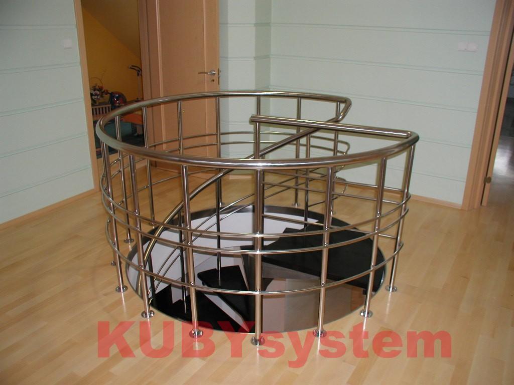 Klatka schodowa z metalowymi, spiralnymi schodami w jasnym wnętrzu. Chromowane barierki, drewniana podłoga. Widoczne drzwi wejściowe i fragment pomieszczenia na piętrze.