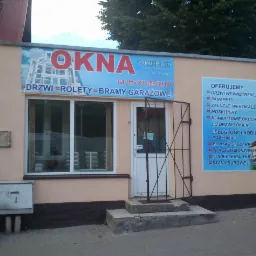 Fasada budynku firmy Zamber-Bud w Giżycku z reklamą okien, drzwi, rolet i bram garażowych; widoczne okno wystawowe i wejście zabezpieczone kratą.