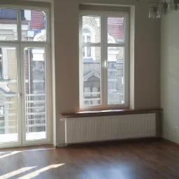 Jasne wnętrze z dwoma oknami z widokiem na budynki. Pod oknem grzejnik panelowy. Na podłodze panele imitujące drewno. Minimalistyczny wystrój.