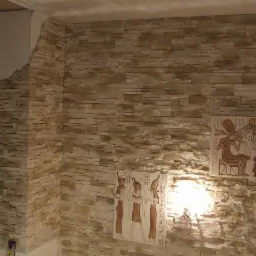 Ściana wykończona kamieniem dekoracyjnym w odcieniach beżu, z egipskimi płaskorzeźbami i oświetleniem punktowym, tworząca unikalny, tematyczny wystr&oacute;j wnętrza.