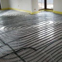 Ułożone rury ogrzewania podłogowego na folii z siatką w jasnym wnętrzu, przygotowane do zalania posadzką, widoczne okna i taśmy zabezpieczające ściany.