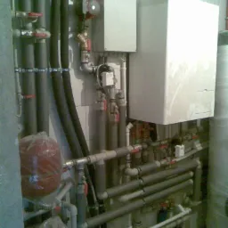 Złożona instalacja z rur, zaworów i urządzeń grzewczych, w tym biały piec, zamontowana na ścianie z jasnymi płytkami. Widoczne izolowane rury i elementy sterujące.